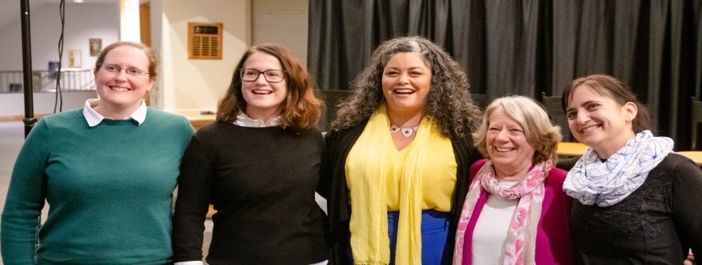 Five women stand before a black curtain in a theater lobby with their arms around each others backs smiling before a group panel discussion in a theater lobby