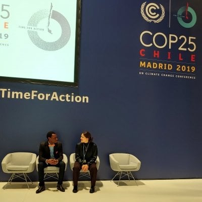 A man and a woman, both seated, looked at each other while smiling. They sit underneath a banner that reads "COP25 Chile Madrid 2019 UN Climate Change Conference".