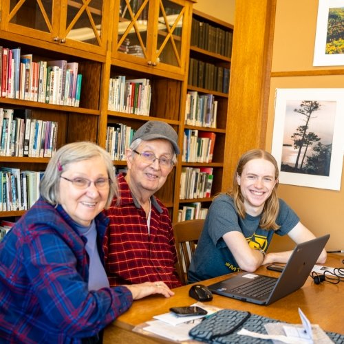 An older couple and a younger man with a Michigan Tech Huskies shirt on smile at the camera in a library.