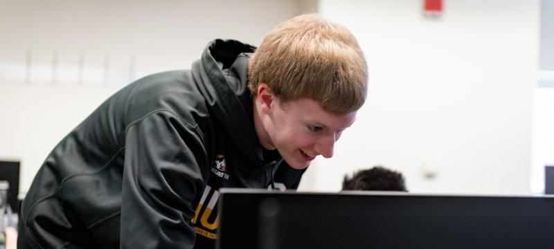 a young man in a black and gold husky sweatshirt leans toward a computer screen