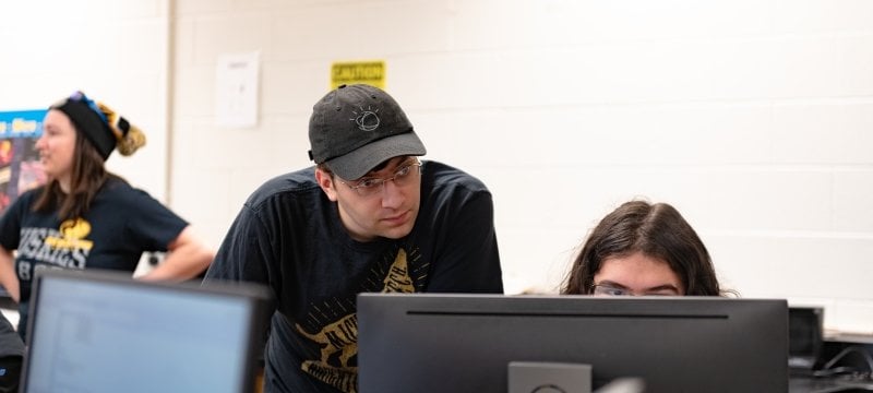 two Michigan Tech students watch students on computer monitors in a lab, one of the students, a girl, is looking intently at the screen