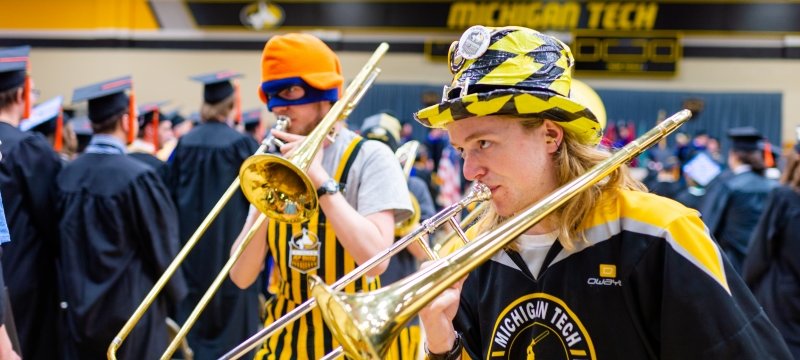 two young men in black and gold stripes play horns in front of black-gowned graduates in a wood gym