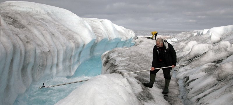 A man in the foreground of a melting glacier with blue water holds a long camera boom into the ice melt as another man stands in the background on an ice sheet.