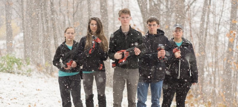 Five students stand in the snowy woods as snow falls from the sky holding power tools and wearing safety glasses