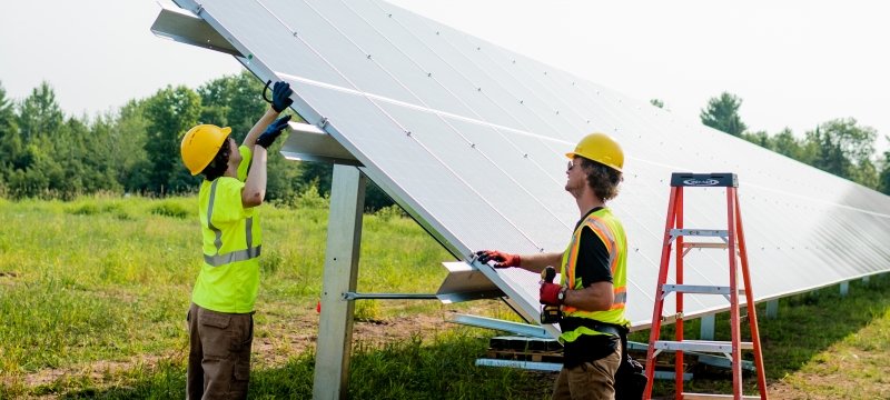Two men wearing construction vests and hard hats attach a solar panel to its footing.