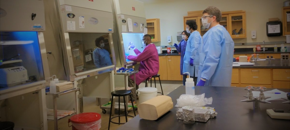 people standing apart from each in a lab with face masks, gloves, and lab coats