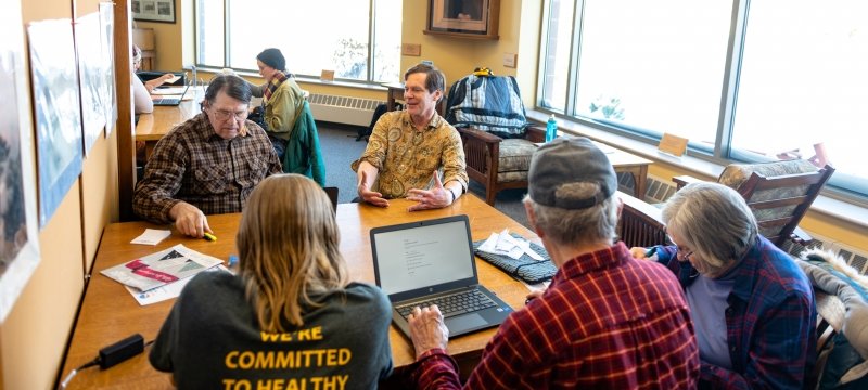 People in a library sitting at tables with a group in the foreground with three people facing away from the camera, one has the word healthy on his shirt, and two older men facing the camera, with sun coming in through the windows and two more people at a table in the background with books on shelves.