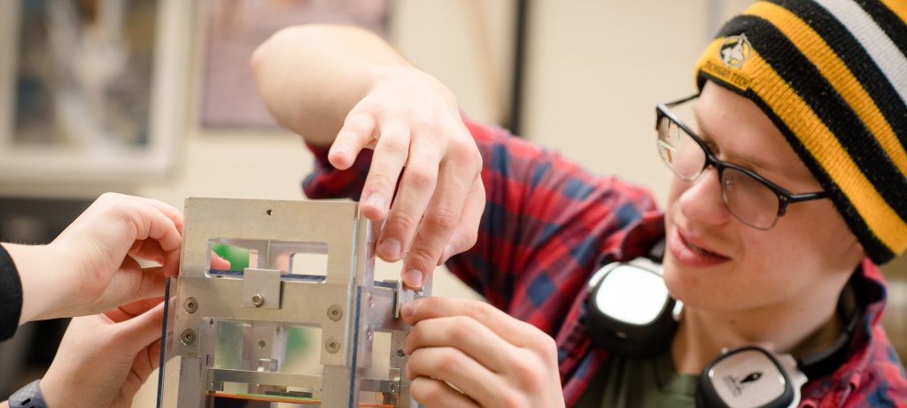 a young man in a knit gold and black striped hat with the Michigan Tech Husky logo on it works on a metal cube which is a nanosatellite being assembled