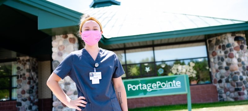 a young woman with a pink face mask on wearing scrubs in front of PortagePointe elder nursing home in Hancock, Michigan