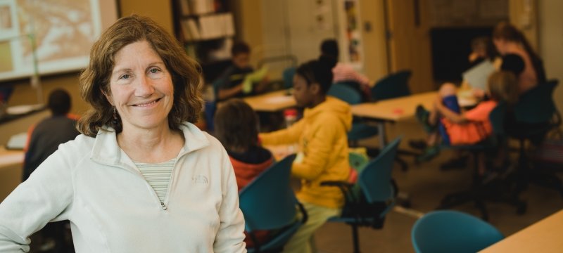 A woman smiles in a classroom with young children seated at desks in the background in a STEM education and environmental outreach program.