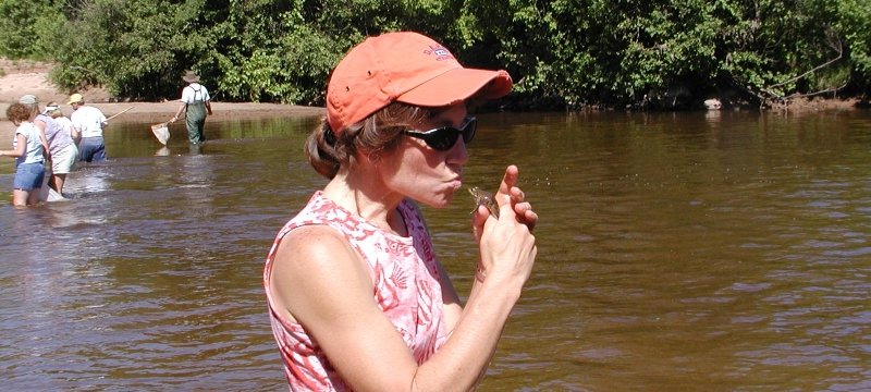 a woman standing in a creek kisses a frog and people with nets and sampling buckets are in the background outside in the summer.