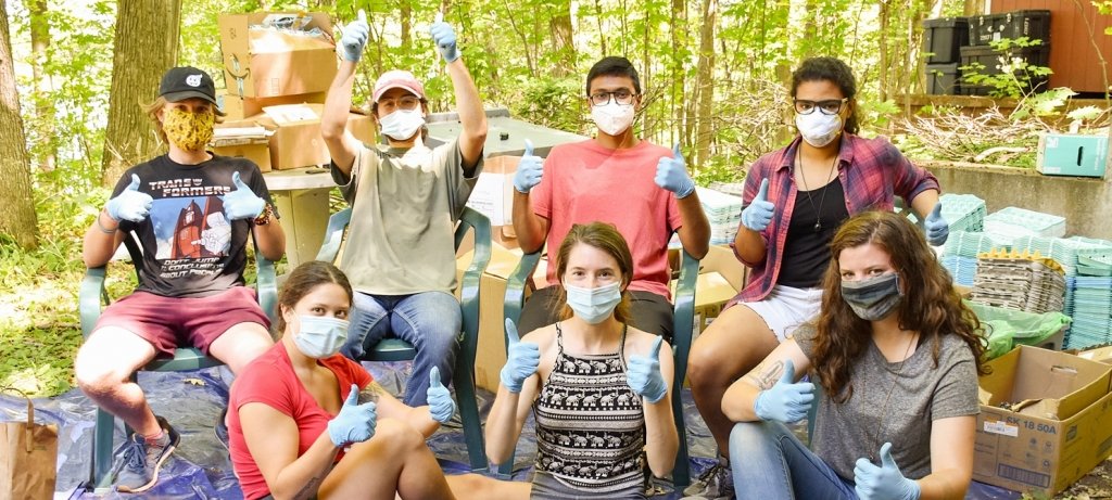 A group of eight students with COVID-19 masks on sitting outside in the woods on a tarp with a brick house behind them and egg cartons and boxes around them. (Photo by Ranit Karmakar)