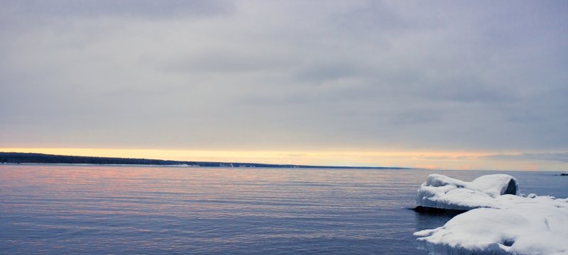 Keweenaw Bay with ice-covered rocks in the foreground of the image and a sunrise just beginning to peak through the clouds.