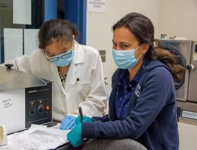 Two women review lab protocol documents. They are wearing face coverings. One is looking at the camera and smiling.