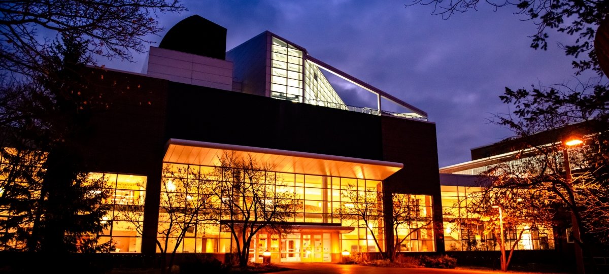 Michigan Tech alum Cathy Leslie learned tenacity during her studies at Michigan Tech. The Dow building on the Michigan Tech campus glows with light just before dawn. The sky is somewhat cloudy in the background and the building is surrounded by trees.