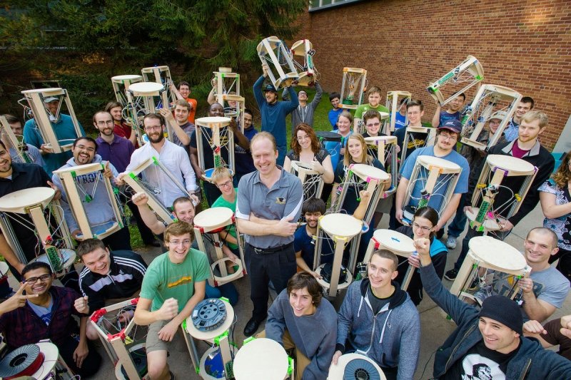 kids with 3D printers and a professor outside with a brick building and tree in the background