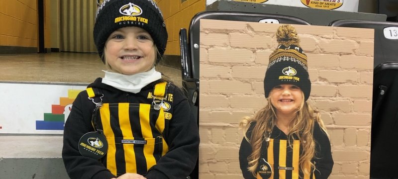 A little group with a Michigan Tech Huskies hat and black and gold striped overalls sits with her fan cutout sign with her Michigan Tech hat on.