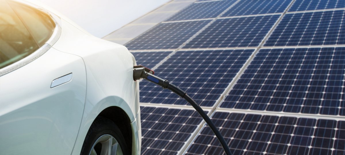 An electric vehicle that is plugged in has a solar panel behind it.