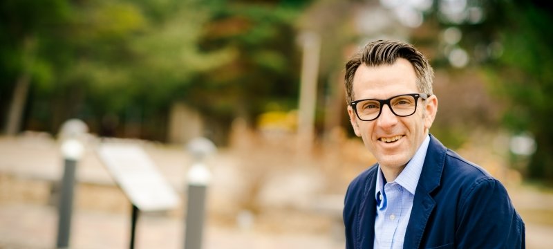 A man wearing glasses and a dress shirt and jacket smiles at the camera seated outside on a college campus with walkway lights and trees in soft focus behind him