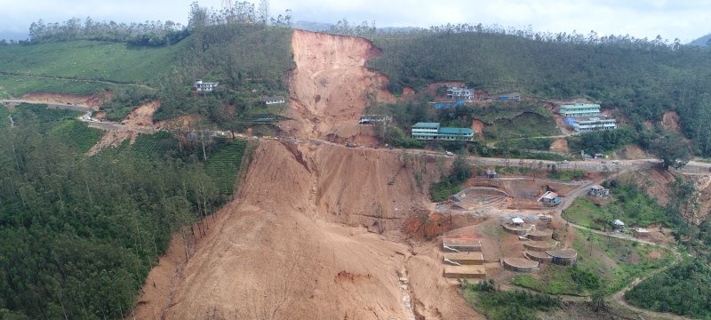 A debris flow washed out a road and covered buildings; the rest of the land is covered in trees. 