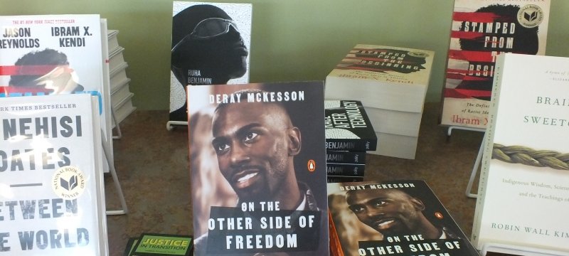 books in stands and stacked on a library table with a green wall in background: from left, counterclockwise, Ta-Nahesi Coates, Between the World and Me; On the Other Side of Freedom, DeRay McKesson; Braiding Sweetgrass, Robin Wall Kimmerer; Stamped from the Beginning, Ibrim X. Kendi, Ruha Benjamin book cover with black woman's head and glasses.   