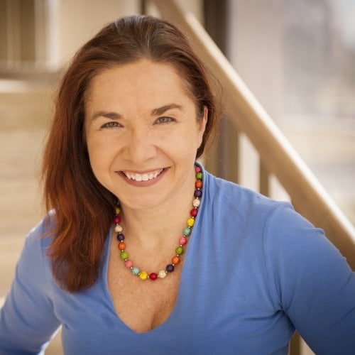 A woman with long hair and colored bead necklace smiles at the camera with a stairway in the background.