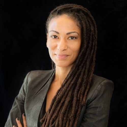 A woman with long braided hair looks at the camera with her arms crossed and a black background.