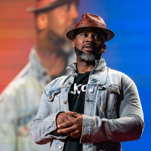 a man in a fedora wearing a headset on a stage at a TED talk with his image projected on a screen behind him.