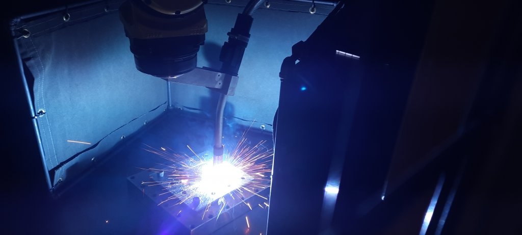 A MIG robot prints a metal part as sparks fly from the tip of the welder on a black background.