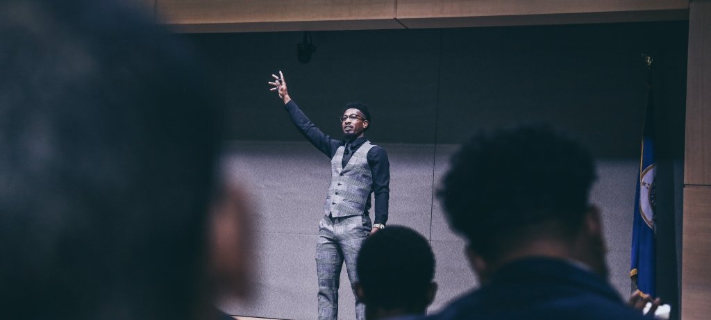 A young man with arm upraised stands on a stage speaking to a crowd at Michigan Tech for MLK day. We can see the back of the audience's heads in the foreground.