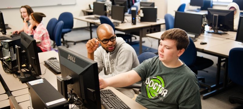 four people in a computer lab at a university working on computers. Older students are teaching youngsters to code.