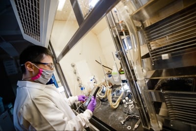 A person in a white lab coat wearing protective eyewear and a cloth face covering uses a handheld instrument while watching a chemical reaction under a laboratory fume hood.