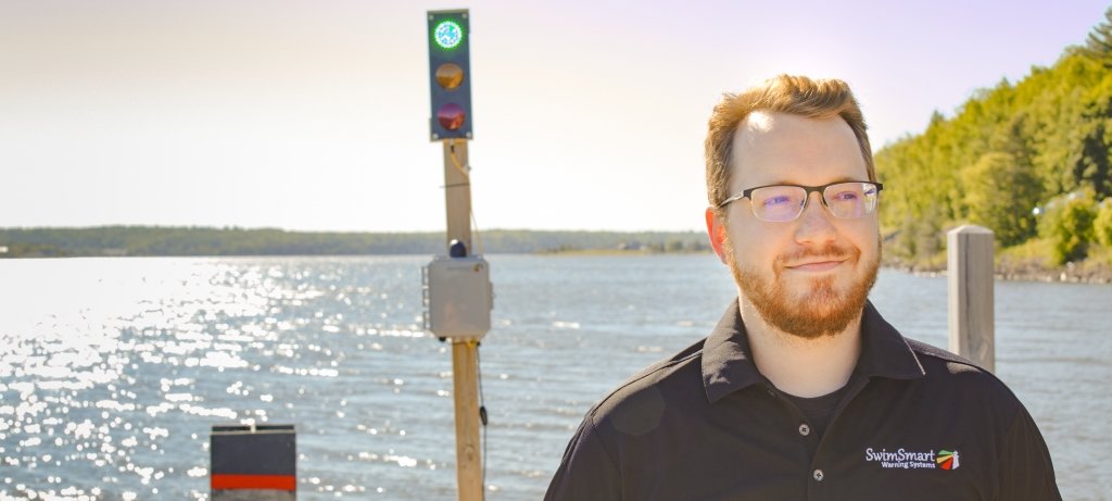 A young man wearing glasses gazes off into the distance with a pole with a green light on it behind him like a traffic signal on a dock on the Keweenaw waterway on the Michigan Tech Campus. His polo shirt reads SwimSmart Warning Systems.