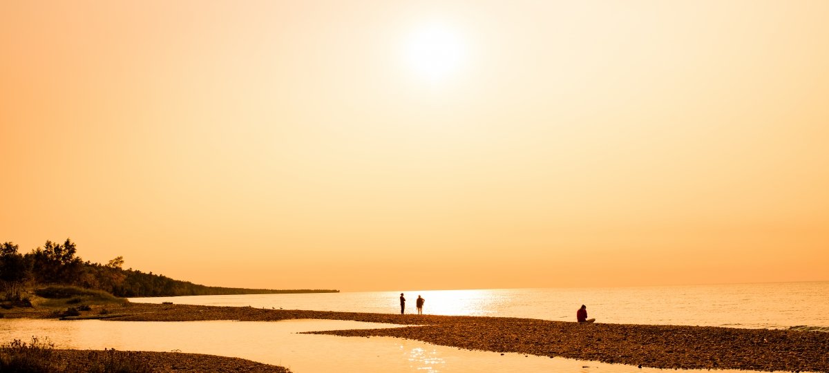 The sun shines over a beach. The sky and the water appear to be the same color. Some people walk on the beach in the distance.