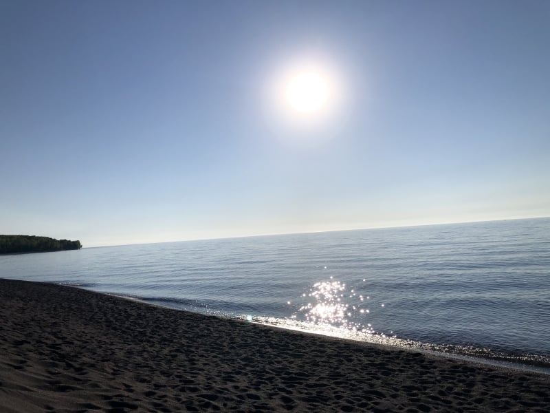 The sun shines over a rocky beach with the tree-lined shoreline in the distance.