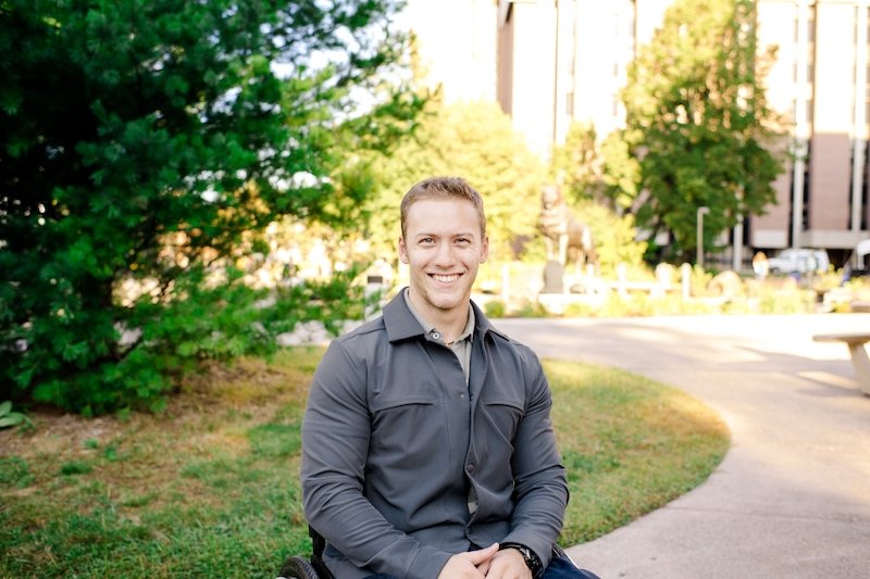 A young man sits smiling behind the Husky Statue on a college campus.