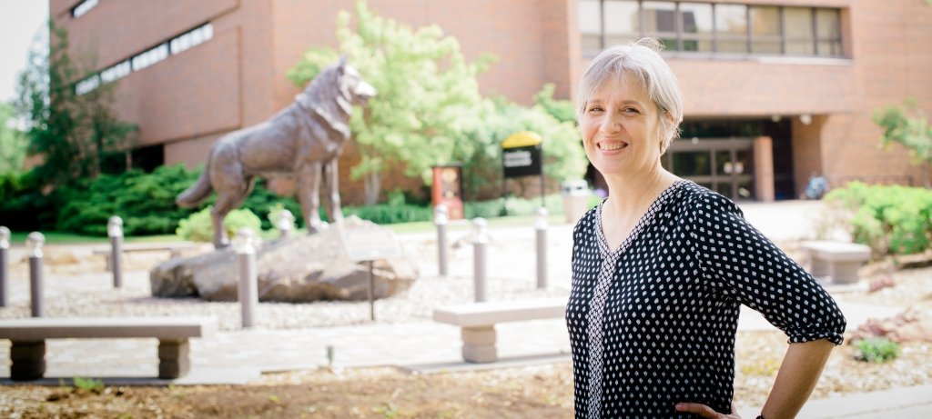 A woman with a hand on her hip smiles with a Husky statue and plaza and a brick building behind her.