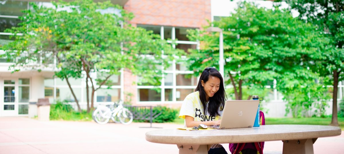 A young woman sits outside at a picnic table and is smiling while working on her laptop.