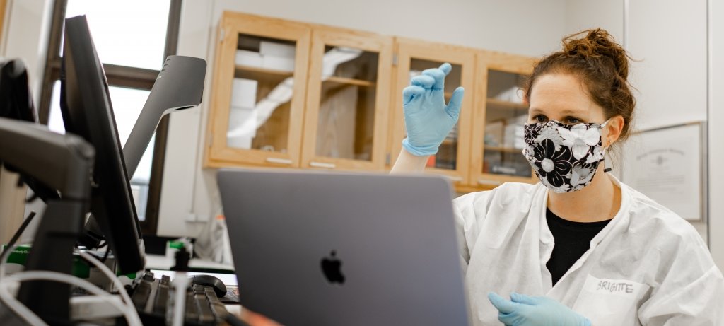Brigitte Morin in the virtual learning lab, encourages students to think critically about their own biases as well as what others bring to the table when navigating difficult dialogues. A woman wearing a face mask and blue gloves gestures to students on a screen in a virtual learning lab on the MTU campus.