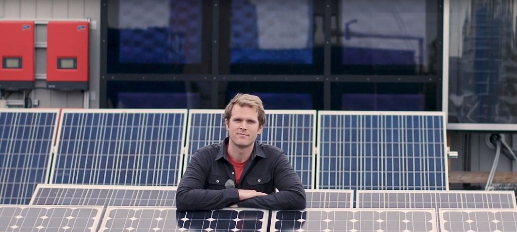 A man leans on a solar panel array with solar panels arrayed behind him and two red control panels in the background.