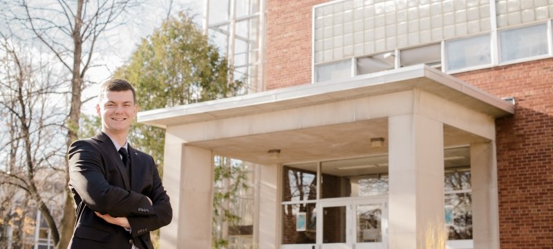 A young man in a suit stands outside in front of a brick and glass building with trees behind him on the Michigan Tech campus.