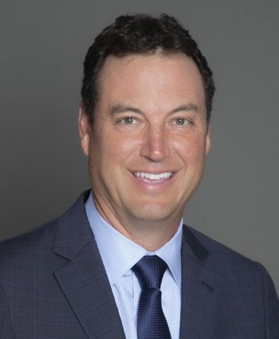 A man smiles in a suit and tie with a neutral plain background.