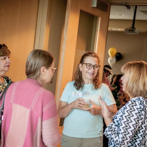 Four women with a background of gold and black balloons at a reception indoors stand talking. Two have their backs to the camera, in daylight.