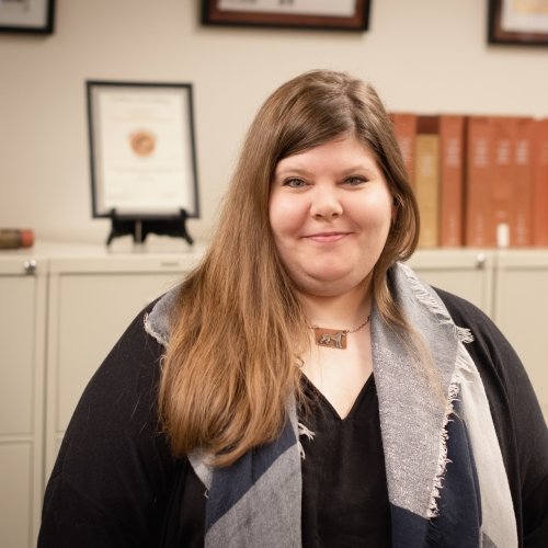 A smiling woman with tall file cabinets and framed certificates behind her.