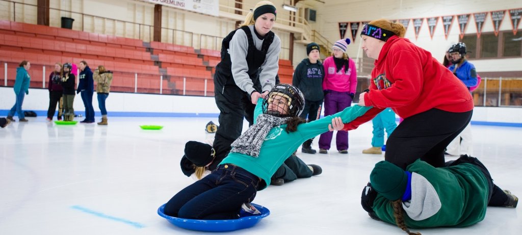 Students on an ice rink prepare to fling a student on a saucer as a human bowling ball as other students watch in the background during a winter carnival competition.