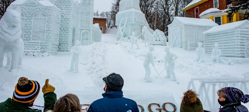 People point at a snow sculpture featuring a giant husky and other snow figures including a shafthouse with a yellow mansion in the background outside in winter.