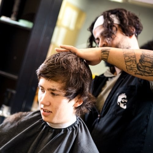 A young man gets his hair barbered wearing a cape with the stylist behind him.