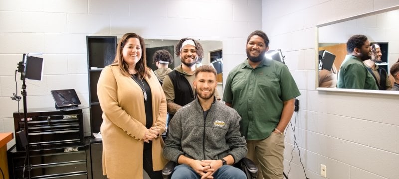 Four people smile, reflected in a mirror, in a pop-up barbershop on Michigan Tech's campus with a mirror reflecting behind them.