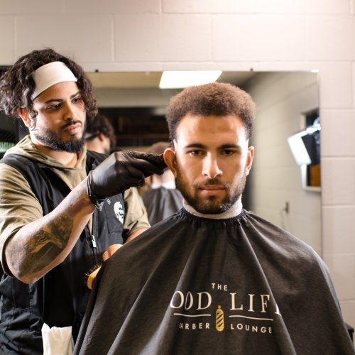 A man gets a haircut in a barber's chair with the barber holding scissors behind him. He wears a cape that says Good Times Barber Lounge.