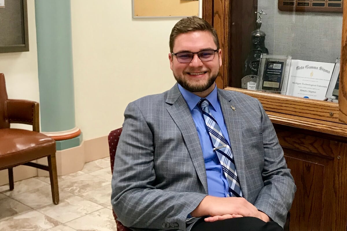 A young man in a tie smiles with a bookshelf behind him in the MTU College of Business